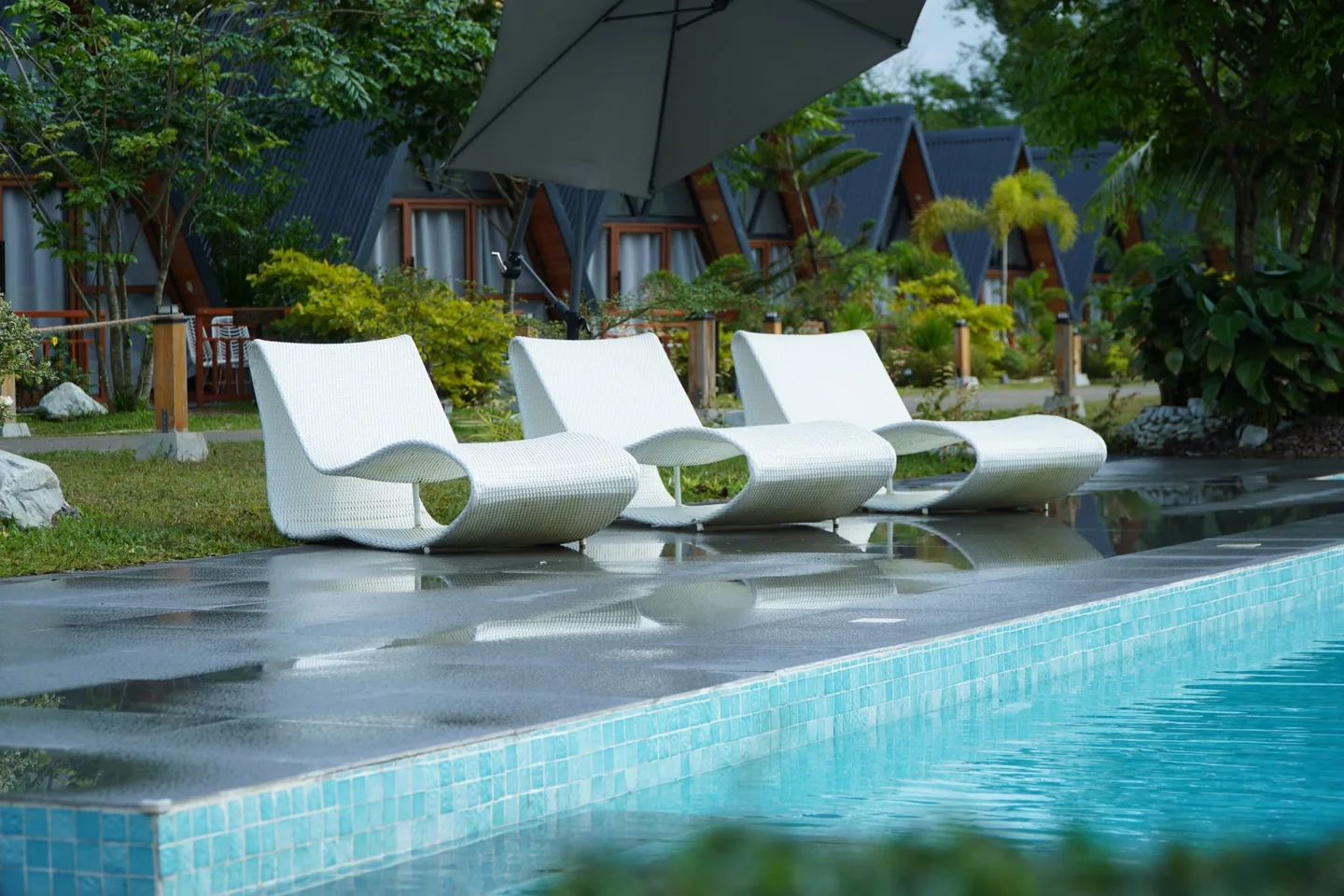 White poolside loungers beneath an umbrella with A-frame cabins beyond